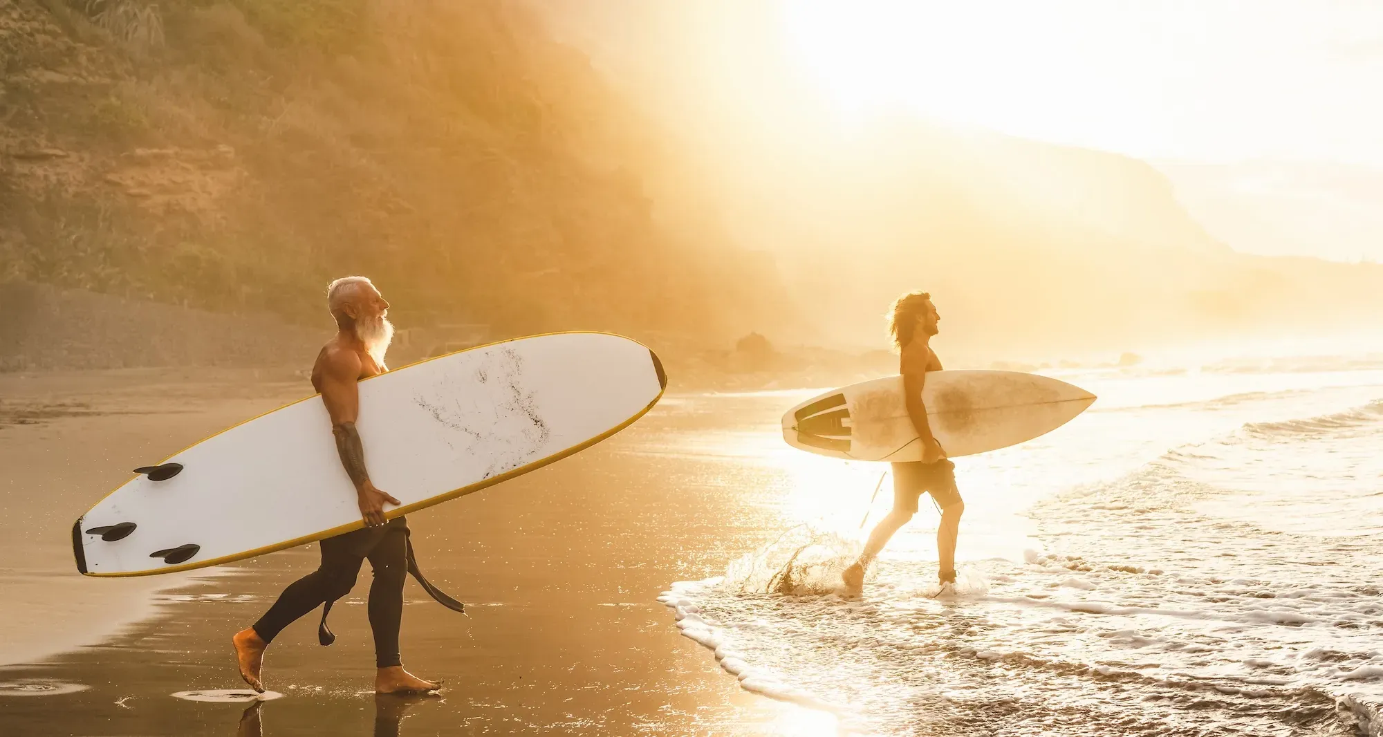 two male surfers on a beach carrying boards heading into the water